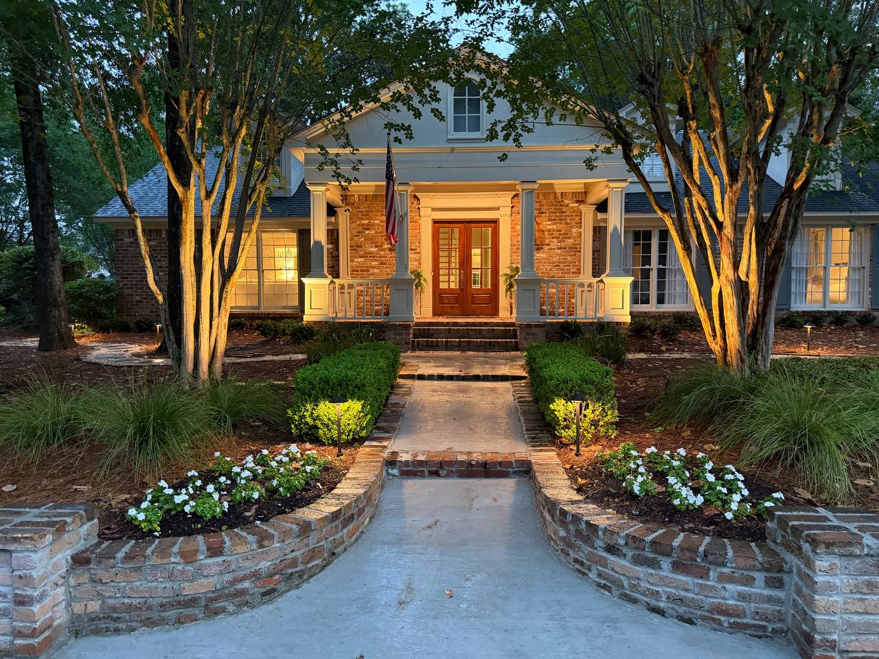 Front entry walk with brick retaining walls, white flowers, and warm lighting leading to a columned brick home at dusk