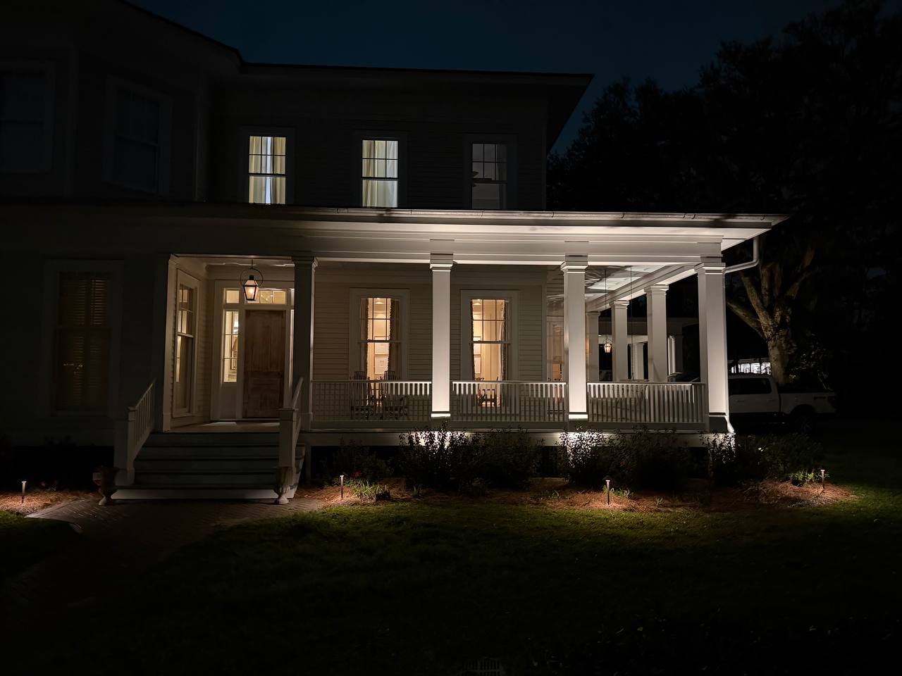 Front-facing night view of a deep porch with evenly lit columns, glowing windows, and soft path lights at the lawn edge