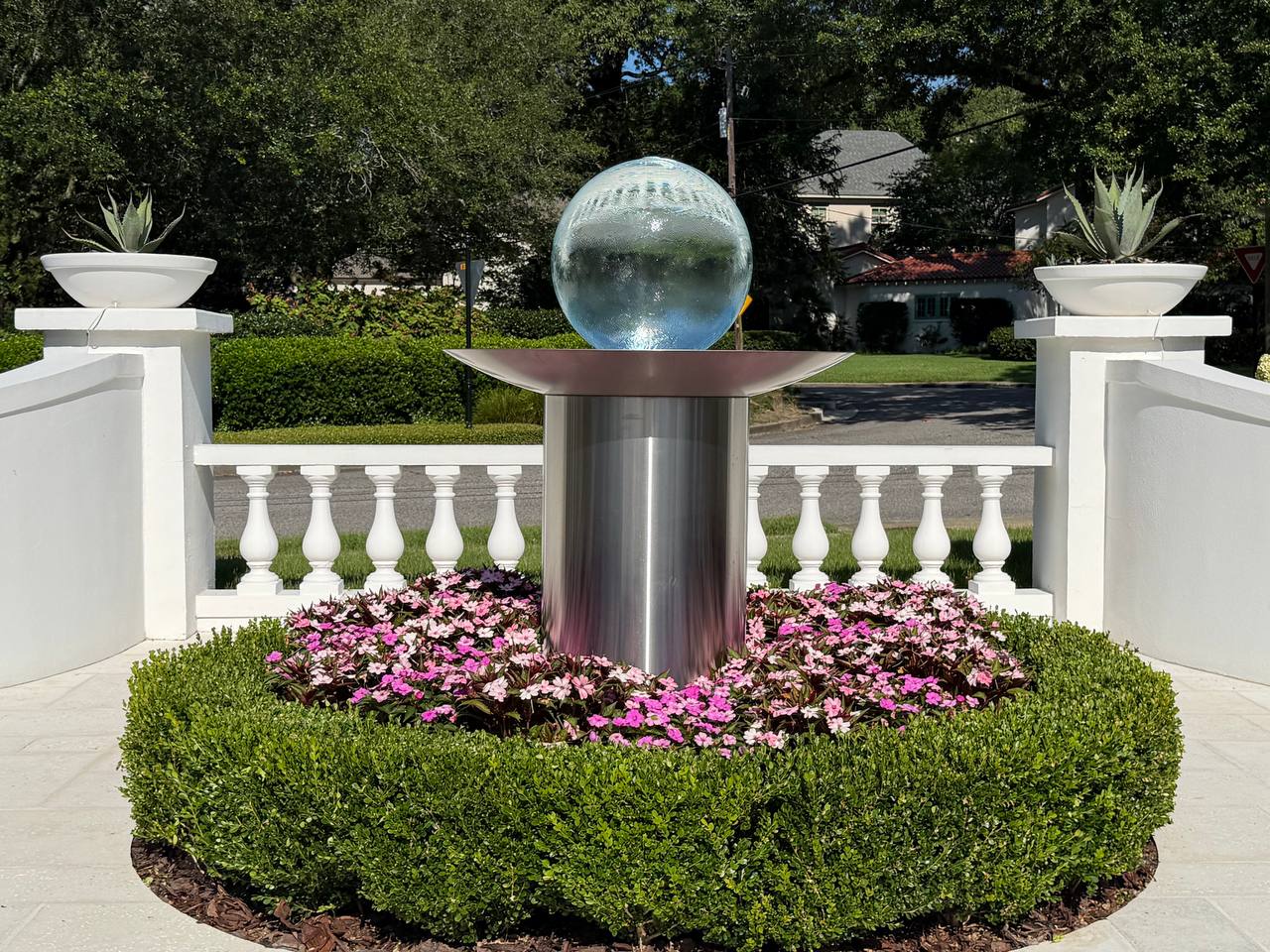 Close detail of a circular boxwood hedge framing pink flowers around a glass globe fountain