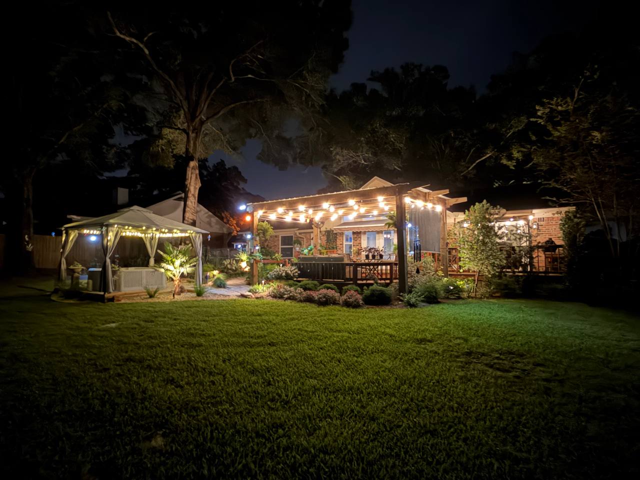 Backyard spa garden with warm lighting across lawn, canopy, planting beds, and patio
