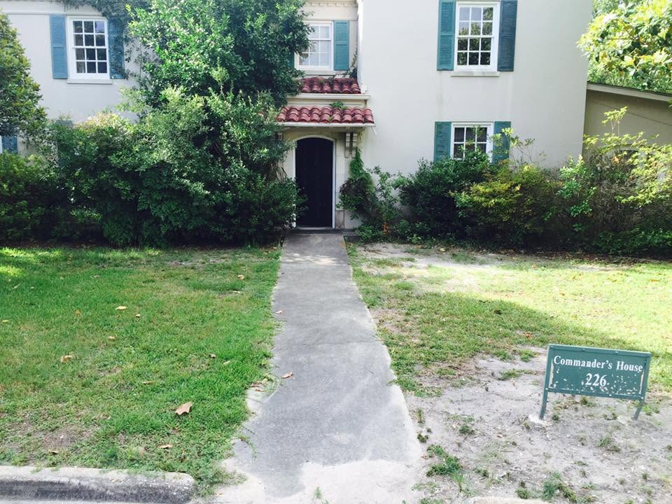 Stucco commander's house before film-preparation landscaping, with sparse front yard, bare soil, and overgrown shrubs near the entry