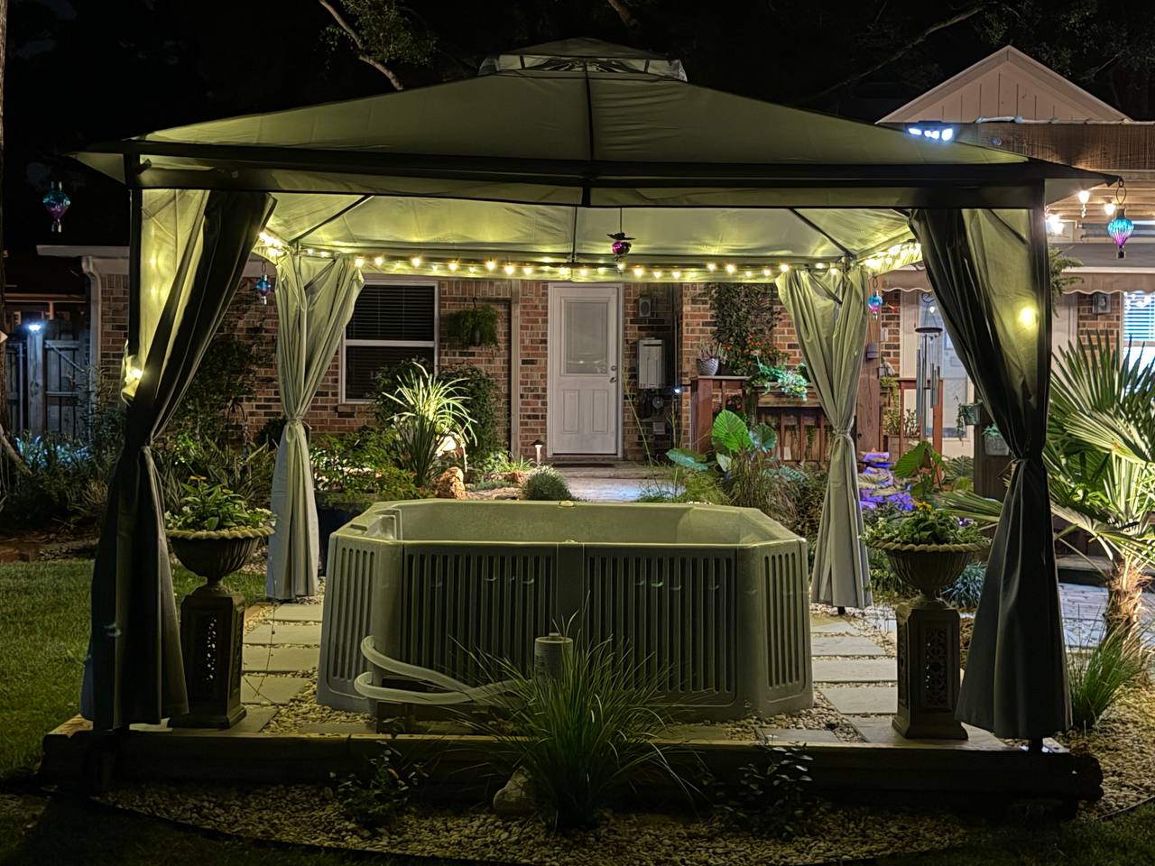 Front-facing spa view beneath a lit canopy with planting beds and stone pavers