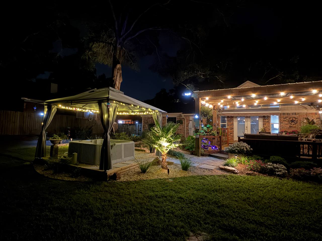 Night view across a backyard lawn toward a spa canopy and warmly lit outdoor living space